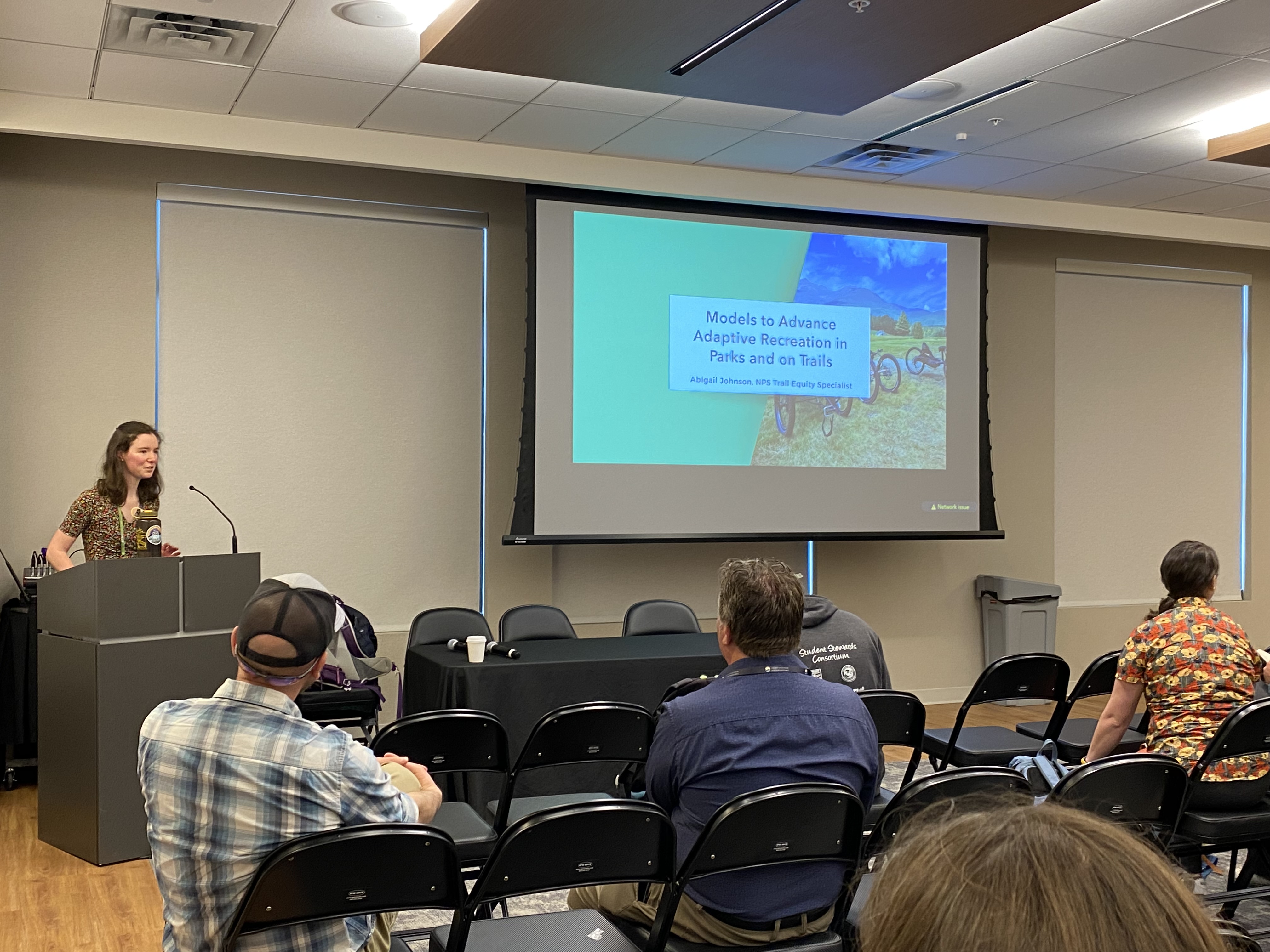 A woman stands behind a podium in front of people in rows of chairs. There is a slide on the screen that says 'Advancing Adaptive Recreation in Parks and on Trails'